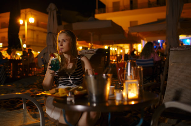 Young woman enjoying a drink in a pub or restaurant sitting at a table alone outdoors illuminated by electric lamps sipping a drink