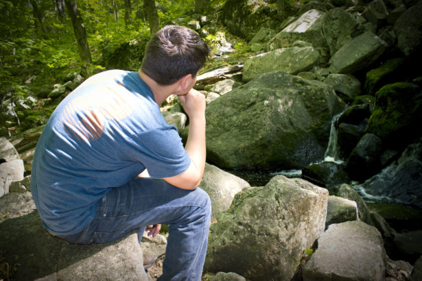 A young man in deep thought while sitting on some rocks near a beautiful waterfall.